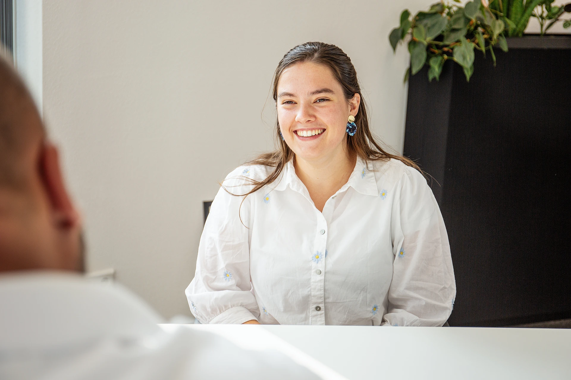 Vrouwelijke collega kijkt lachend in de camera, in gesprek met een andere collega